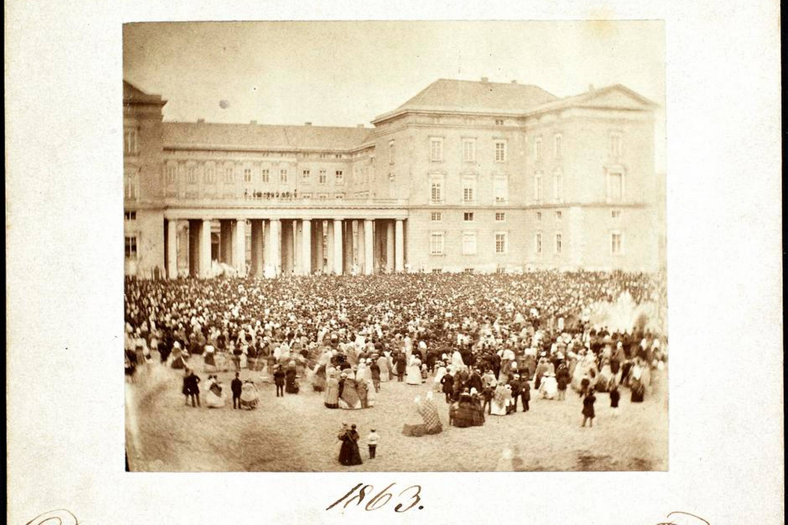 Grundlovsmøde på Christiansborg Ridebane i 1860’erne. Kongen og hans følge står på den søjlebårne balustrade. Den 5. juni blev fra 1849 en national mindedag, og særligt efter forfatningskampens begyndelse i 1870’erne blev det en dag med stærke politiske manifestationer. Også i 1850’erne bølgede ”den første del” af forfatningskampen. Det Kgl. Bibliotek