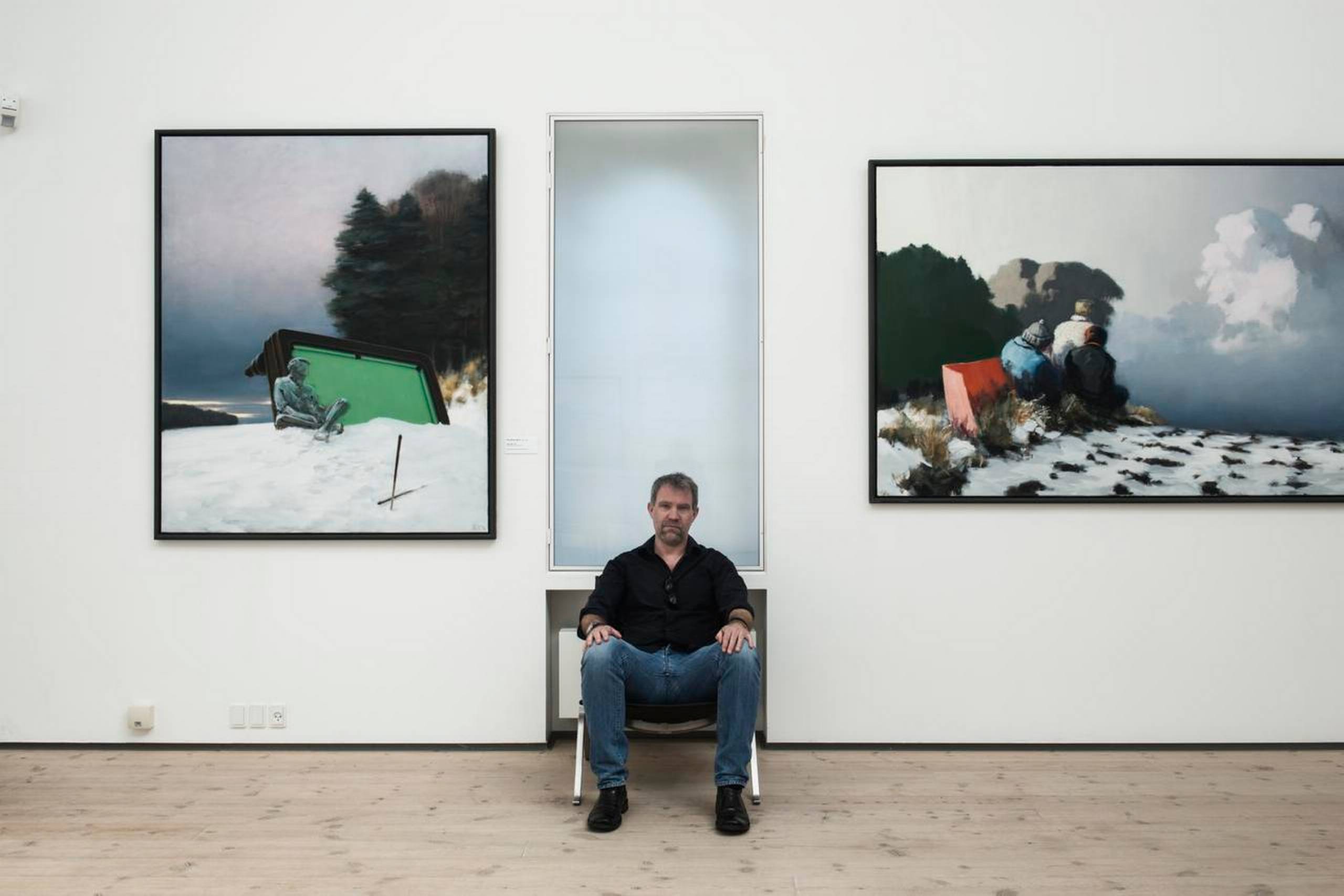 Forfatter Jakob Knudsen sidder mellem to malerier af Poul Anker Bech på Vendsyssel Kunstmuseum. Poul Anker Bech (1942-2009) er kendt for at male minimalistiske billeder, der ofte har et surrealistisk element, fortæller Jakob Knudsen. Han nævner også, at Poul Anker Bech var billedkunstlærer på Hjørring Gymnasium, mens han selv gik der. Foto: Casper Holmenlund Christensen